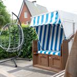 Terrasse mit Strandkorb und Hängesessel. Hintergrund: grüner Heckenzaun und Haus.