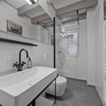 Badezimmer mit Dusche, Waschbecken und Toilette. Weiße Fliesen, schwarze Armatur und goldener Türknauf.