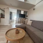 Wohnbereich mit grauem Sofa, runden Tisch und Fernseher. Küche im Hintergrund.