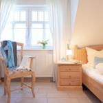 Ein Schlafzimmer mit Holzbett, Nachttisch und Stuhl. Fenster mit Vorhängen und Blumen auf dem Fensterbrett.