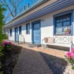 Haus mit blauen Fenstern und Tür, weißer Bank mit Kissen, Blumenbeete und gepflastertem Weg.