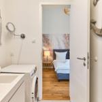 Badezimmer mit Waschmaschine und Blick in Schlafzimmer mit Bett und Holzboden.