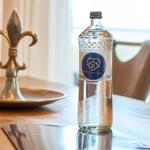 Eine Wasserflasche mit Logo steht auf einem Holztisch neben einem silbernen Fleur-de-Lis-Objekt.