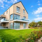 Moderne Villa mit Glasbalcony und grünem Garten unter blauem Himmel.
