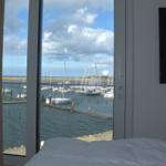 Blick auf Hafen mit Segelbooten durch großes Fenster. Bett und Wandfernseher sichtbar.