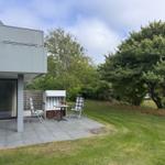 Terrasse mit Gartenhaus, Tisch und zwei Stühlen. Grünfläche und Bäume im Hintergrund.