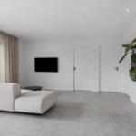 Wohnzimmer mit Sofa, Fernseher und Pflanze. Große Fenster und weiße Wände.