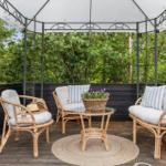 Terrace with pavilion, three rattan chairs, table, and plants. Surrounded by trees and a dark wooden wall.