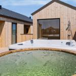 Außenbereich mit Whirlpool und Holzterrasse. Hintergrund: Holzhaus mit Glasfront.