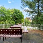 Terrasse mit Tisch und Bänken, umgeben von grünen Bäumen und einem weißen Zaun.