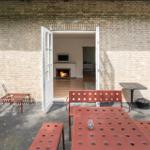 Terrasse mit rotem Metallmöbel und offenem Durchgang zum Innenraum mit Kamin.