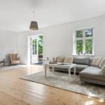 Wohnzimmer mit Sofa, zwei Sesseln, Tisch und Fenster. Holzboden und Tür zum Garten.