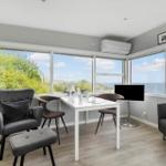 Ein Raum mit Tisch, Stühlen und Fernseher. Große Fenster bieten Blick auf Meer und Bäume.