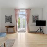 Wohnzimmer mit Holzboden, Sofa, Fernseher auf Stativ und Fenster mit rosa Vorhängen.