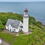 Weißes Haus und Leuchtturm auf grüner Halbinsel am Meer mit Windkraftanlagen im Hintergrund.