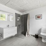 Badezimmer mit Dusche, Waschbecken und Toilette. Weiße Wände, graue Fliesen und Fenster.