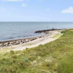 Strand mit Sand und Grasflächen, das Meer und ein kleiner Steinschutz sind sichtbar.