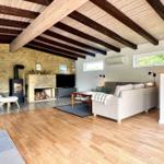 Wohnzimmer mit Holzboden, Kamin und Sofa. Deckenbalken und Fenster sichtbar.