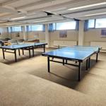 Zwei Tischtennistische in einem Raum mit Fenstern und Sofas.