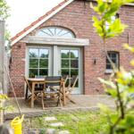 Roter Backsteinbau mit Terrasse und Holztisch. Garten mit Gras und Bäumen.