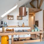 Küche mit Holzinsel, weißen Schränken und Dachfenster. Holzkonstruktion und Küchenutensilien sichtbar.