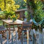 Holztisch mit Stühlen und Sonnenblumen auf Terrasse. Hängemattensitz unter Baum.