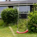 Ein roter Schaukelstuhl hängt im Garten vor einem Haus mit grünen Sträuchern.