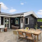 Terrasse mit Tisch, Stühlen und Grill. Schwarze Holzwände und weißer Kamin. Blumen und Pflanzen.