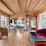 Wohnraum mit Holzdecke, Fenstern und rotem Sofa. Im Hintergrund ist ein Schlafzimmer sichtbar.