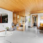 Living area with wooden ceiling, fireplace, and large window. Bookshelf and seating areas.