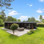 Modern black cabin with wooden terrace and garden. Outdoor seating area under trees.