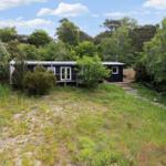 Black wooden house on stilts, surrounded by trees and grassy areas with stone steps.