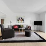 Living room with sofa, armchair, rug, and TV. Large windows with garden view.