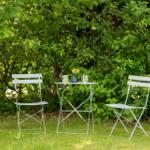 Zwei weiße Stühle und ein Tisch stehen im Gras vor dichtem Grün.