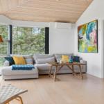 Wohnzimmer mit Sofa, Tisch und Blick auf Wald. Holzdecke und Boden. Wandbild und Kamin.