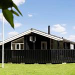 Black wooden house with balcony and grill area on green lawn under blue sky.