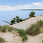 Sandhügel mit Gras, Blick auf ruhiges Wasser und einen Steg.