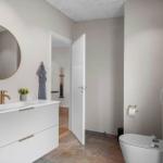 Badezimmer mit Waschbecken, Spiegel und Toilette. Tür führt in angrenzenden Raum.