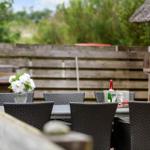 Terrasse mit Tisch, Stühlen, Blumen und Weinflasche. Hintergrund: Holzzaun und Backsteinhaus.
