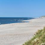 Weiterer Strandabschnitt mit Sand, Meer und Dünen. Einige Personen sind am Wasser.