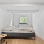 Bedroom with bed, small table, and window. Wooden floor and white walls.