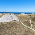 Dune landscape with hiking trail and view of the sea under blue sky.