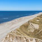 Aerial view of a sandy beach next to dunes and the blue sea.