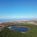 A circular lake is surrounded by a forest, hills, and the sea.