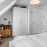 Schlafzimmer mit Dachschrägen, weißer Kleiderschrank, Holzregal und Bett mit weißer Bettwäsche.