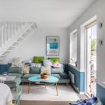 Wohnzimmer mit Sofa, Tisch und Treppe. Wandfarbe weiß und blau. Fenster mit Blick nach draußen.