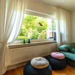 Ein Raum mit grünem Sofa, bunten Sitzkissen und großem Fenster mit Blick auf einen Garten.