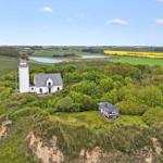 Wohnhaus und Leuchtturm auf grüner Anhöhe mit Blick auf Felder und Wasser.