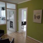 Wohnbereich mit grüner Wand, Sitzgelegenheiten und Blick in Küche und Esszimmer.