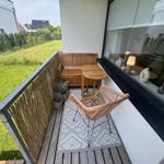 Terrasse mit Holzbank, Tisch und Rattanstuhl. Grünfläche im Hintergrund.